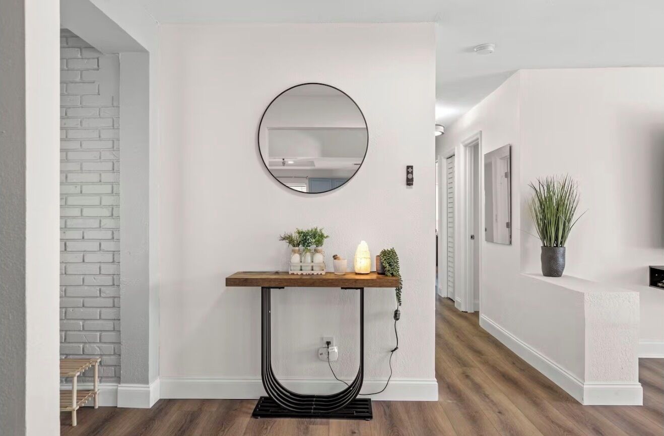 Un couloir avec un mur en briques blanches, un miroir rond, une console en bois ornée d’objets décoratifs et une plante en pot.
