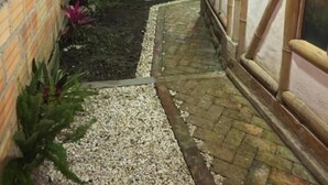 Un estrecho sendero exterior conduce a un área de estar techada, con una mesa y sillas, rodeada de muros de ladrillo y plantas.
