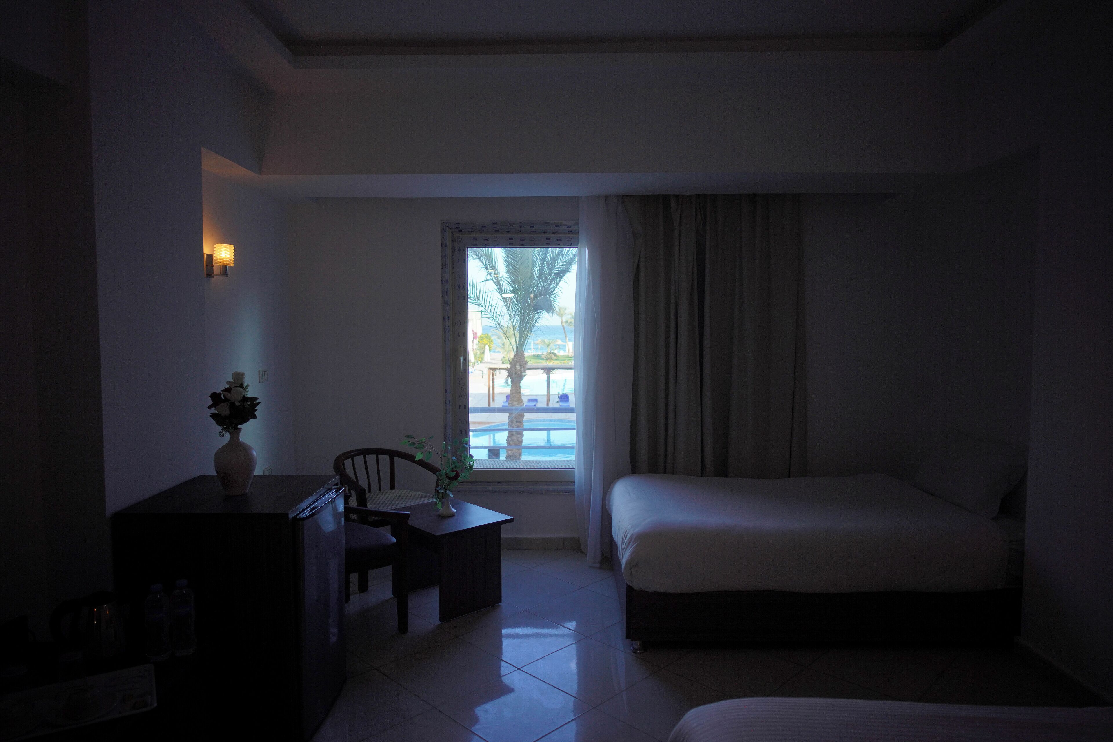 A hotel room with a bed, a desk with a chair, a small table with a vase, and a window with curtains.