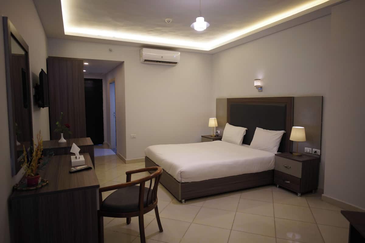 A modern hotel room with a large bed, bedside tables with lamps, a desk with a chair, and an air conditioning unit.