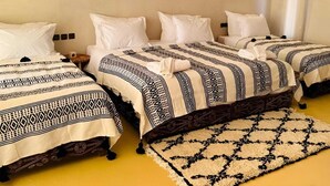Een hotelkamer met drie bedden, elk met beddengoed met een patroon, een ronde spiegel aan de muur en een klein tafeltje met een lamp.