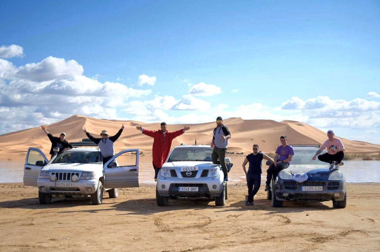 A group of people posing with four cars in a desert setting.