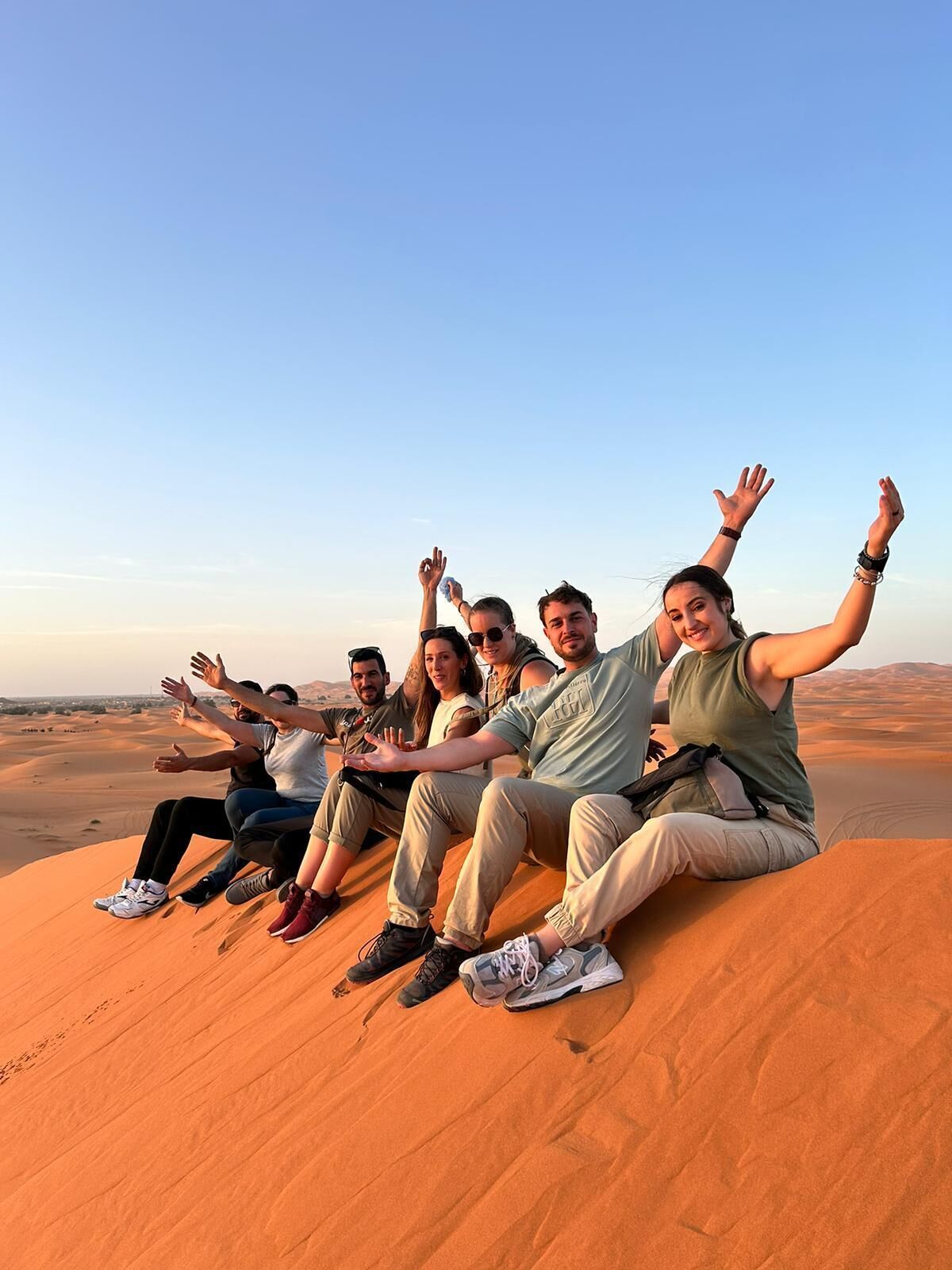 A group of people sitting on a sand dune with a clear sky in the background.