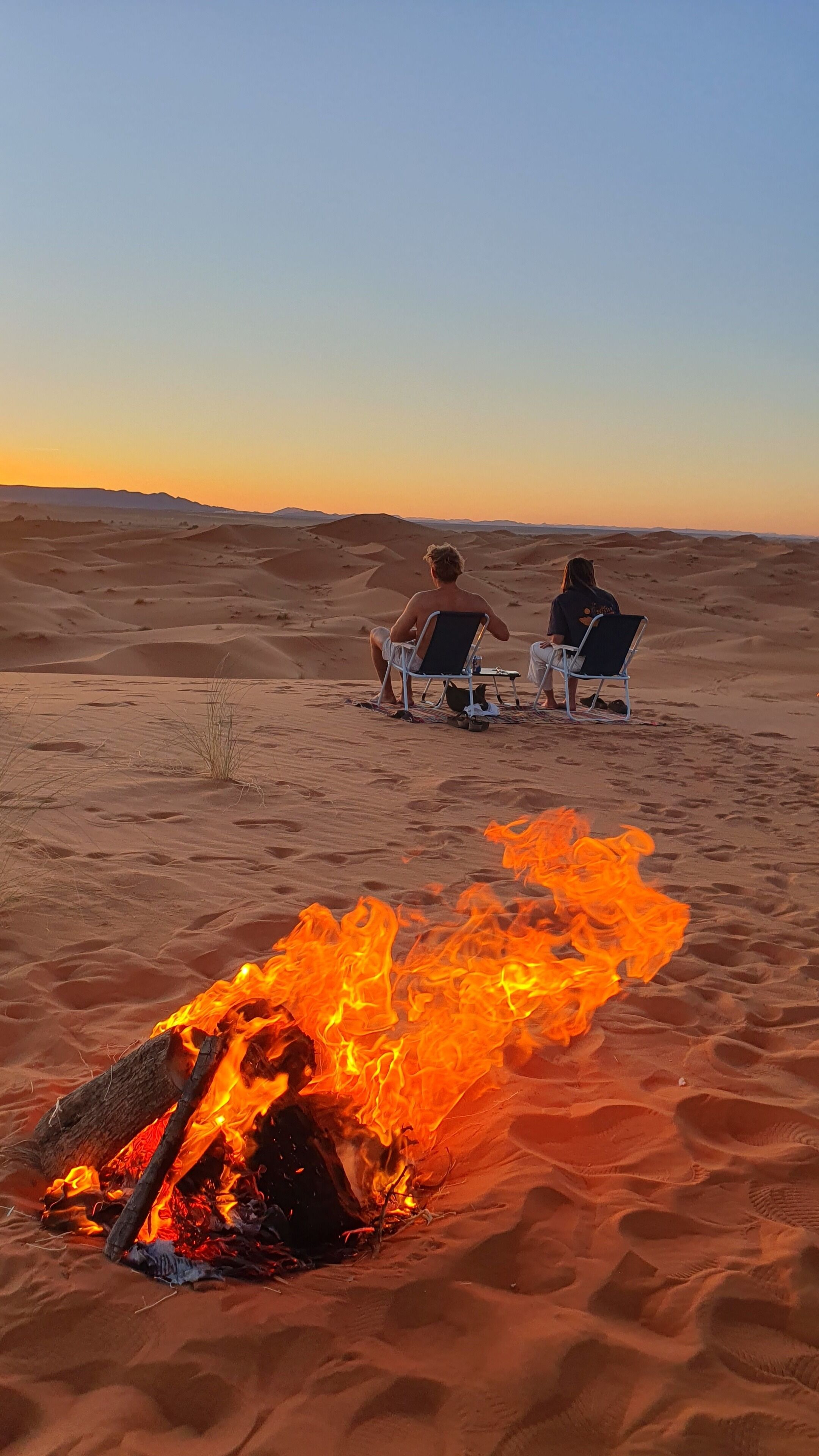 Dos personas sentadas en sillas en un paisaje desértico, con un fuego al frente.