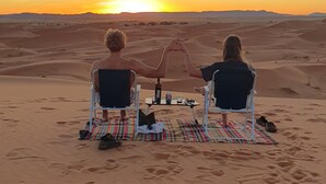 Twee mensen zitten op stoelen en kijken naar een ondergaande zon boven een woestijnlandschap.