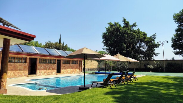 Ein Pool mit Liegestühlen und Sonnenschirmen, ein Steinhaus mit rotem Dach und eine gepflegte Rasenfläche.