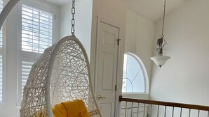 Una silla colgante de mimbre blanco con un cojín amarillo en una habitación con paredes blancas, suelo negro y barandilla blanca.