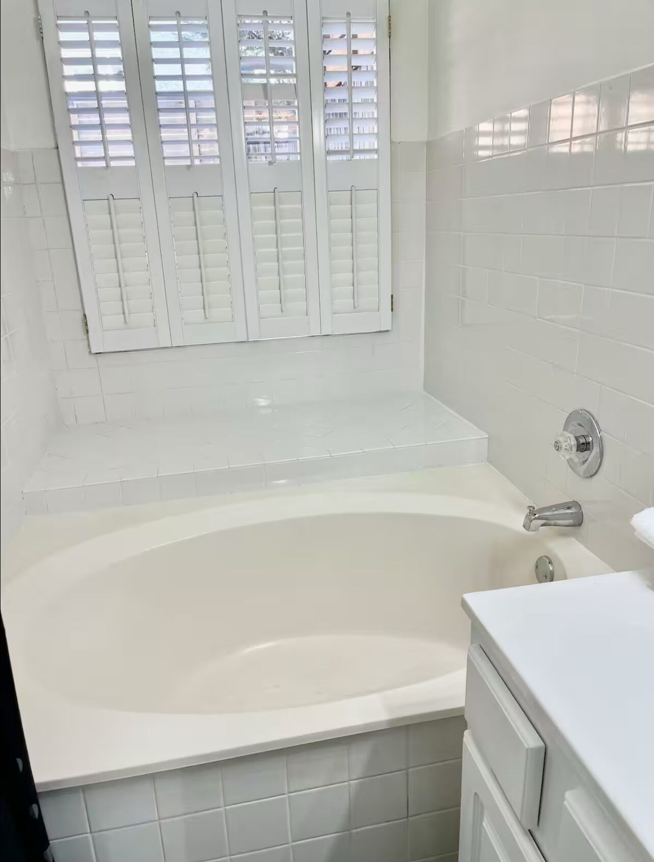Un bagno con una vasca, pareti piastrellate in bianco e una finestra con persiane.