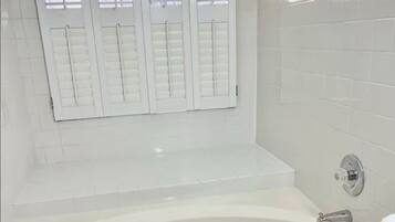 Un bagno con una vasca, pareti piastrellate in bianco e una finestra con persiane.