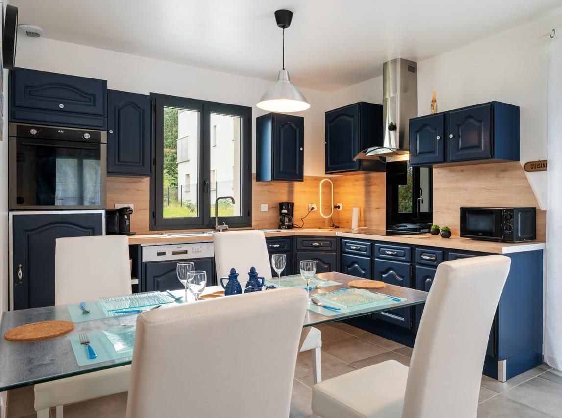 Una cocina moderna con armarios azul oscuro, una isla central y un comedor con sillas blancas y una mesa de vidrio.