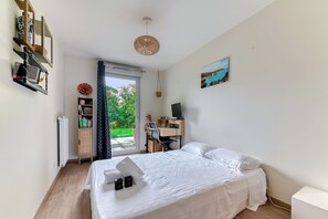 Ein Schlafzimmer mit Bett, Schreibtisch, Stuhl, Computer und einem Fenster mit Blick auf Grünflächen.