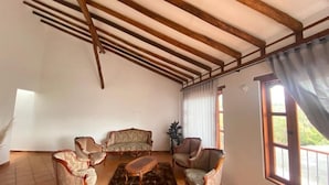 Een woonkamer met een hoge plafond, houten balken, een bank, fauteuils en een salontafel.
