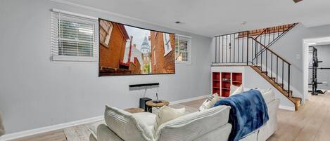 A modern living room with a large flat-screen TV mounted on the wall, a staircase leading to an upper level, and a gym area visible through an open door.