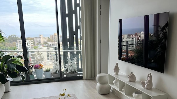 A modern living room with a large window, a white sofa, a coffee table with a vase of flowers, and a mounted TV. - Luxury Apartment with City Views in the Heart of Bondi Junction (Bondi Junction)