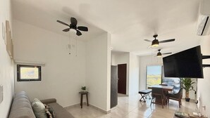 A modern living room with a sofa, a coffee table, and a TV. There are ceiling fans, a dining area with chairs, and a small side table with a plant.