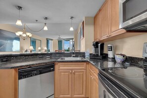 A modern kitchen with a dishwasher, stove, and microwave. It features wooden cabinets, a granite countertop, and pendant lighting.