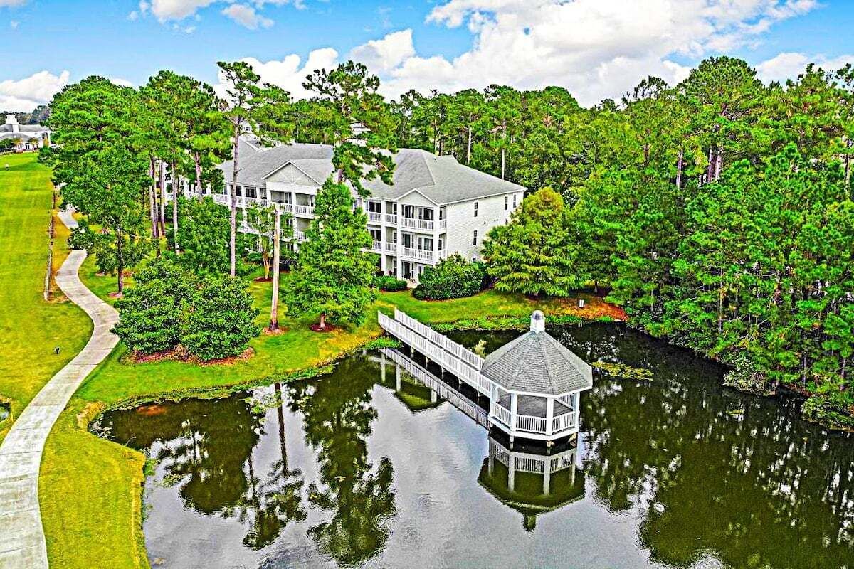 A large, white, two-story house with a gazebo over a pond, surrounded by lush greenery and a golf course.