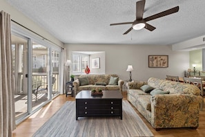 A spacious living room with a ceiling fan, a large sofa set, a coffee table, and a dining area with a table and chairs. - Secluded Condo Near Pool and close to the Ocean! (Myrtle Beach)