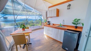 A modern kitchen with a wooden countertop, a sink, and a refrigerator. There is a dining table with chairs and a potted plant. The kitchen has a view of the outdoors through a large window. - Tocuma Glamping (San Rafael)