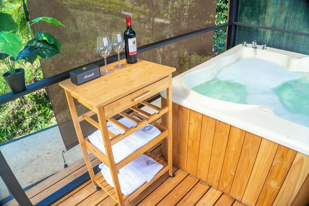 Una mesa de madera con una botella de vino, copas y un altavoz portátil en un balcón con jacuzzi.