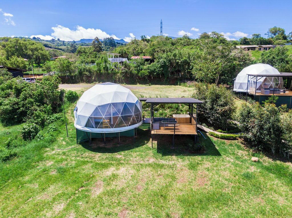 Une structure en forme de dôme, dotée d’une terrasse en bois et d’un espace de repos abrité, entourée d’une végétation luxuriante et d’autres structures similaires dans un cadre rural.