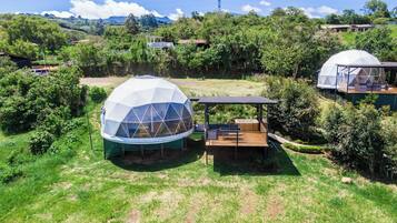 Une structure en forme de dôme, dotée d’une terrasse en bois et d’un espace de repos abrité, entourée d’une végétation luxuriante et d’autres structures similaires dans un cadre rural.