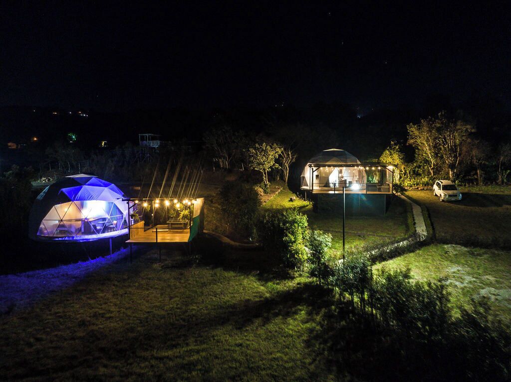 Una estructura en forma de cúpula con fachada de vidrio y terraza de madera, rodeada por un jardín ajardinado de noche.