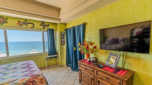 Ein Zimmer mit einem Bett, einem Fernseher, einem Holzkasten, einem Fenster mit Vorhängen und Meerblick.