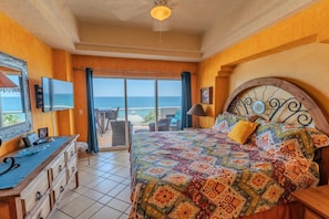 A bedroom with a bed, a TV, a dresser, and a balcony with a view of the sea.