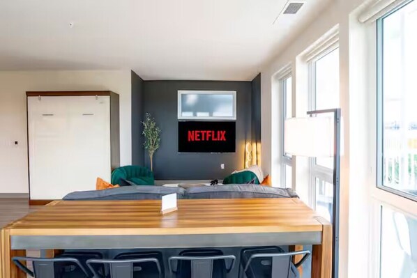 A modern living room with a wooden dining table, a sofa, and a wall-mounted TV displaying "NETFLIX".
