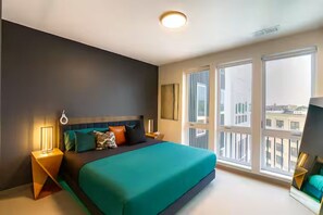 A modern bedroom with a large bed, a nightstand, a lamp, and a view of the city through large windows.