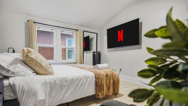 A modern hotel room with a bed, a TV mounted on the wall, and a window with curtains. - 2 BR 1 Bath Apt with Parking | Close to Amenities (Oshawa)