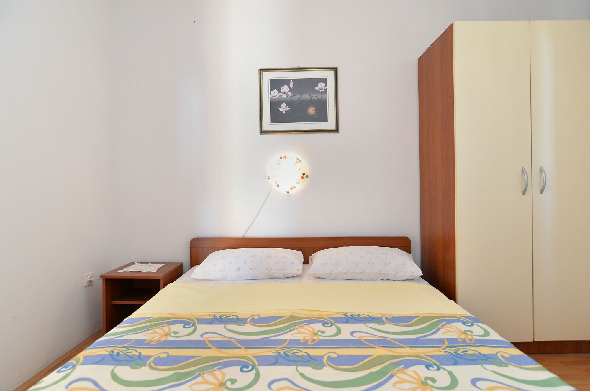 Ein Schlafzimmer mit einem Bett, einem Nachttisch, einem Kleiderschrank, einer Wandleuchte und einem gerahmten Bild.