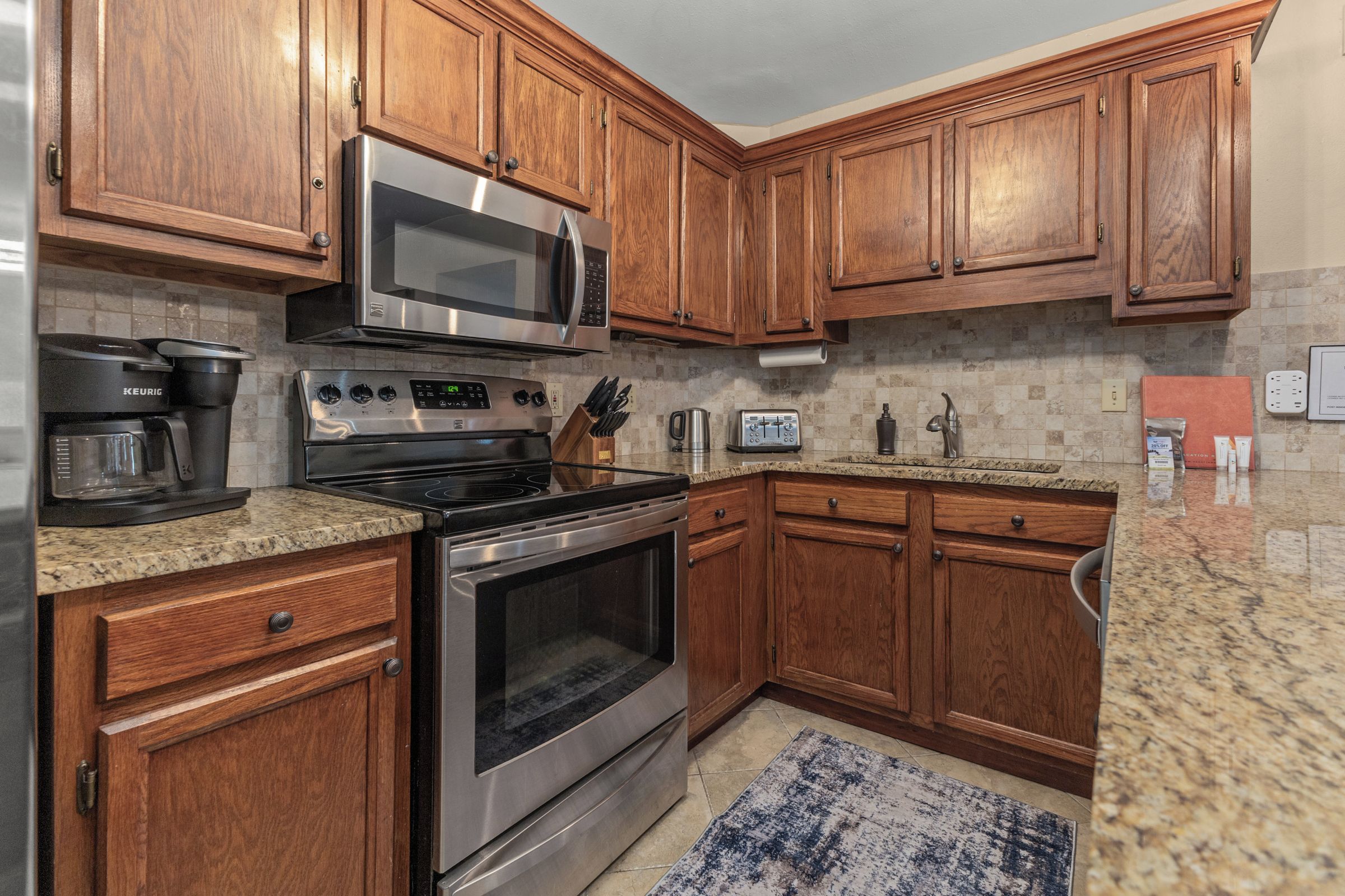 A kitchen with wooden cabinets, stainless steel appliances, granite countertops, and a patterned rug.