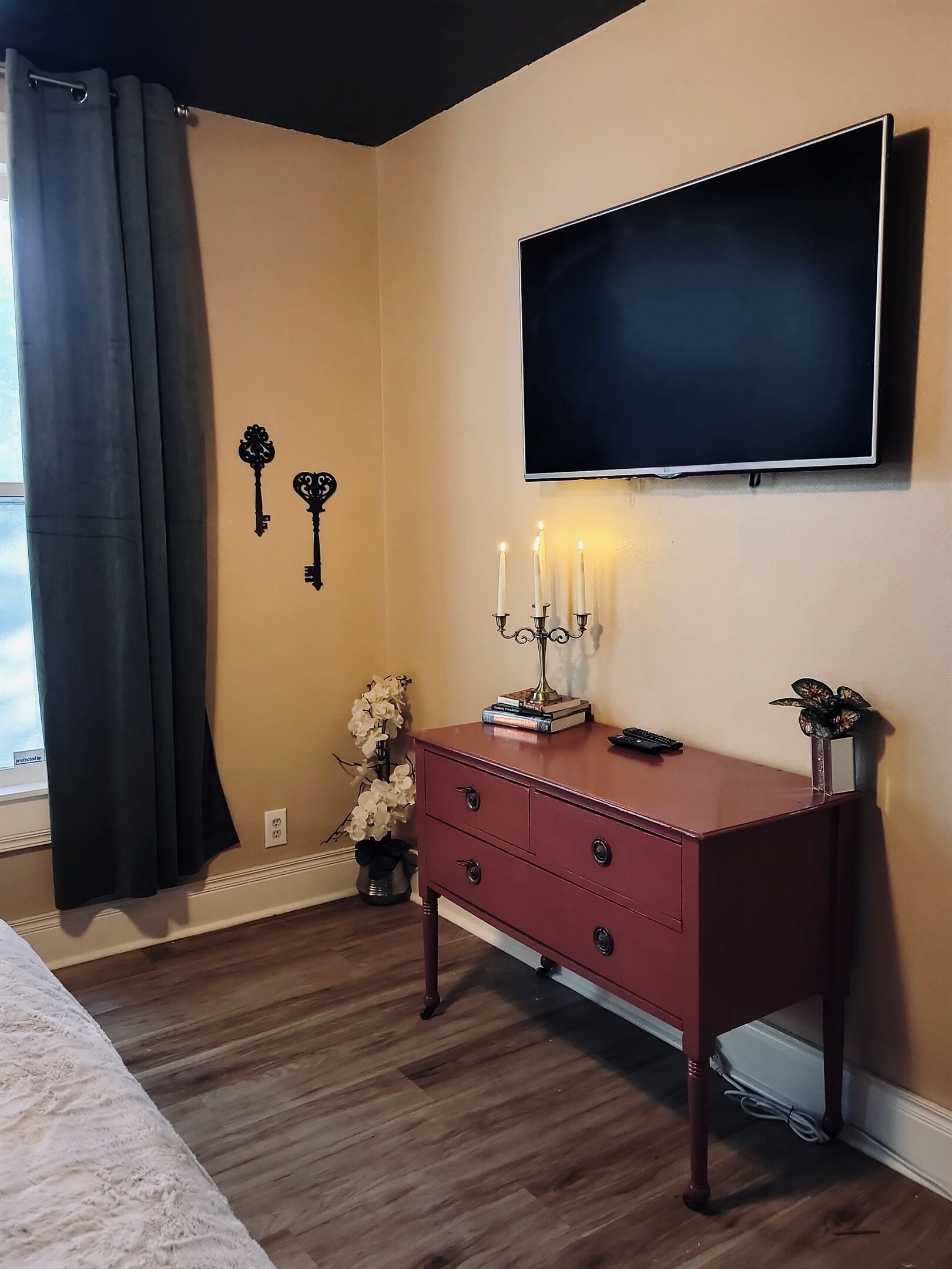Een kamer met een aan de muur bevestigde televisie, een rode dressoir met laden, een vaas met bloemen en een zwart gordijn.
