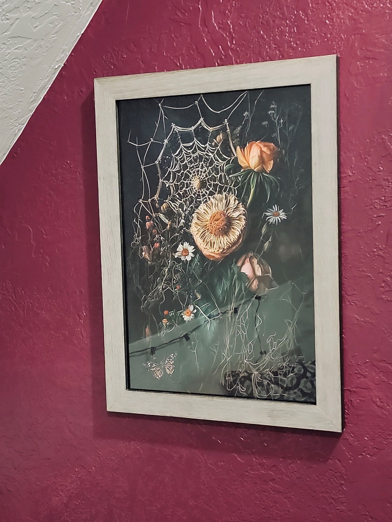 A framed artwork of a spider web with flowers and a butterfly on a textured wall.