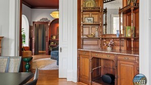 Een ruime woonkamer met een houten kast, een eethoek met een tafel en stoelen, en een gang die naar een andere kamer leidt.
