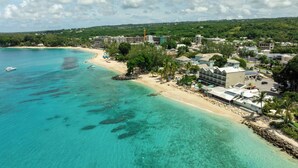 Eine Vogelperspektive zeigt einen Küstenort mit einem Sandstrand, klarem türkisblauem Wasser und einer Mischung aus Wohn- und Hotelgebäuden.
