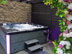 Ein Whirlpool mit Holzbank und Treppen, umgeben von Zierpflanzen und einer strukturierten Wand.