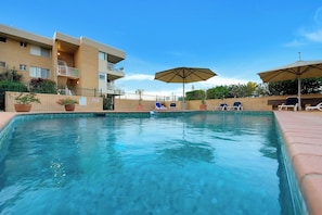 A swimming pool with lounge chairs and umbrellas, surrounded by a brick building with balconies. - Beachfront Escape, Ocean Views, Walk Everywhere (Alexandra Headland)