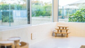 Eine Küche mit weißer Arbeitsplatte, einem Holzschemel und einem Fenster mit Blick auf Grünflächen.