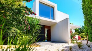 Una casa moderna con un ventanal grande, una terraza de madera y abundante vegetación.