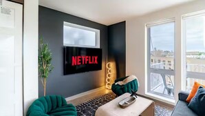 A modern living room with a large window, a flat-screen TV mounted on the wall, and a green sofa. - Modern 5th Floor Sky Suite Retreat - 1BR Sleeps 6 (Milwaukee)