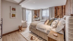 A bedroom with a large bed, a desk, and a chair. There are curtains, a lamp, and a small stool. - Luxury Catered Ski Chalet Blackcomb in Val d'Isere, France LVDI-4 (Val d'Isere)