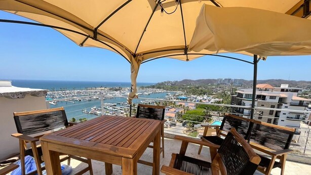 A wooden table and chairs under a large umbrella on a balcony overlooking a marina with boats and buildings in the distance. - Beautiful beachfront condo, 5 min walk from downtown, (Cruz de Huanacaxtle)