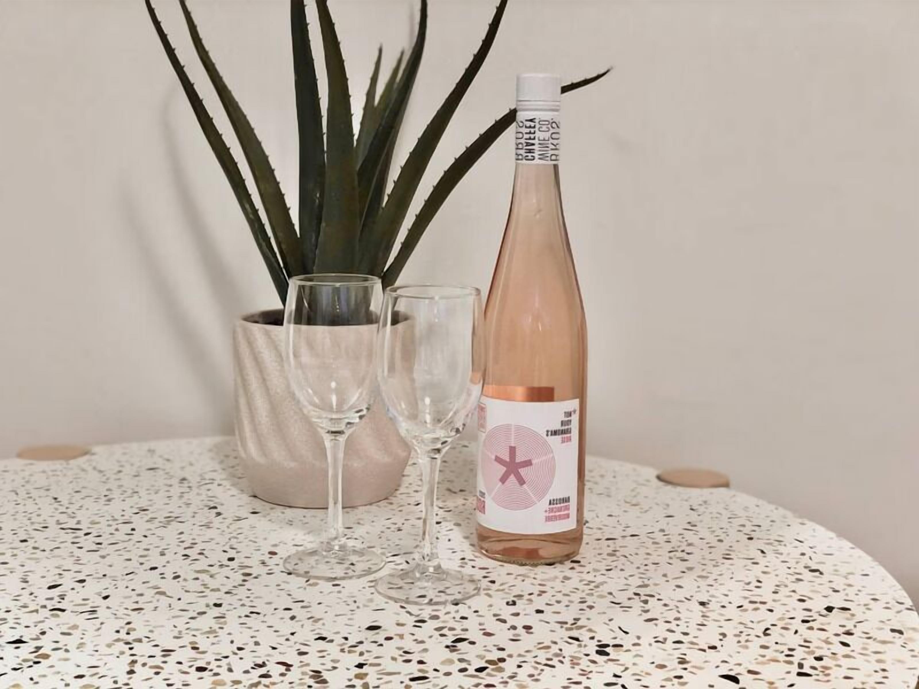 A bottle of rosé wine, two empty wine glasses, and a potted plant on a speckled table.