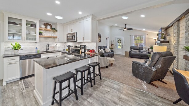 A modern kitchen with white cabinets, a central island with stools, a dining area with a sofa and chairs, and a living room with a fireplace.