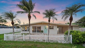 Une maison de plain-pied avec une clôture en bois peinte en blanc, des palmiers et une pelouse bien entretenue.