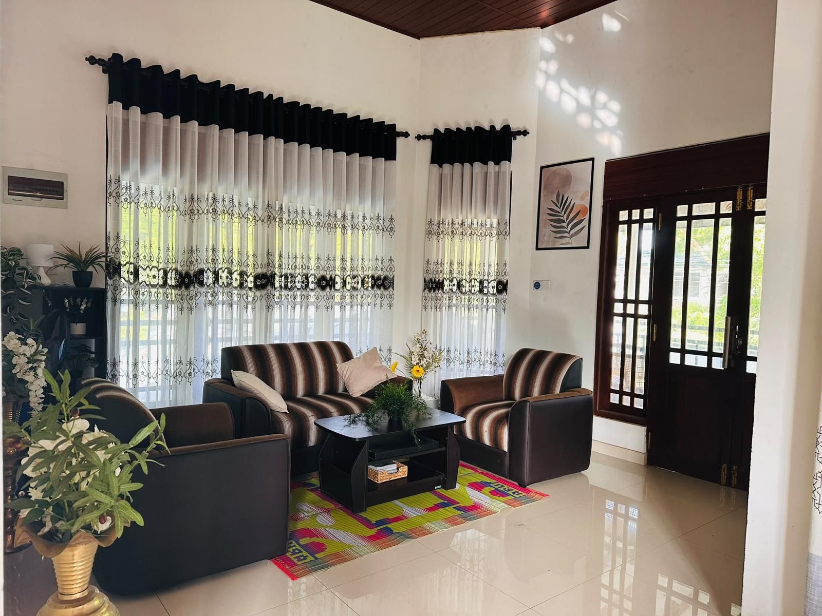 A living room with a sofa set, a coffee table, and a patterned rug. There are two windows with curtains, a door, and a wall-mounted air conditioning unit.