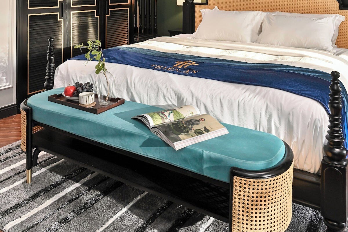 Un dormitorio con cama, mesita de noche con bandeja, banco, ventilador de techo y una ventana grande con cortinas.